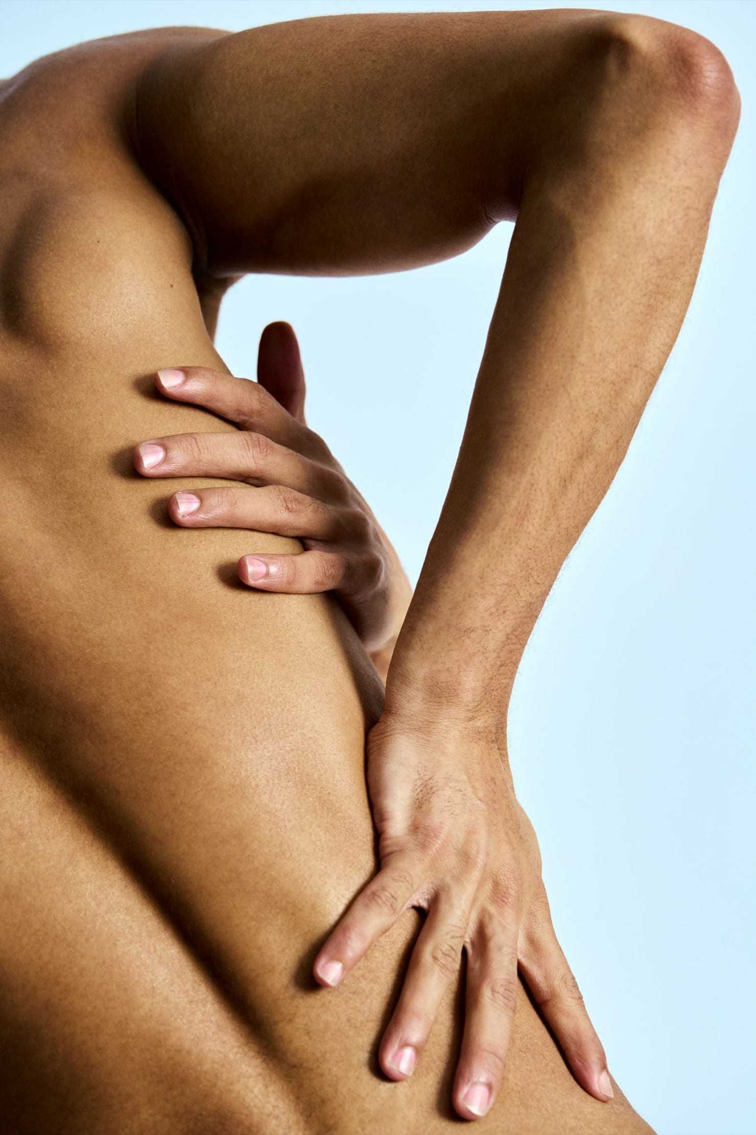 Close-up study of a person's torso and back with hands resting on hips, lit against a soft blue backdrop.