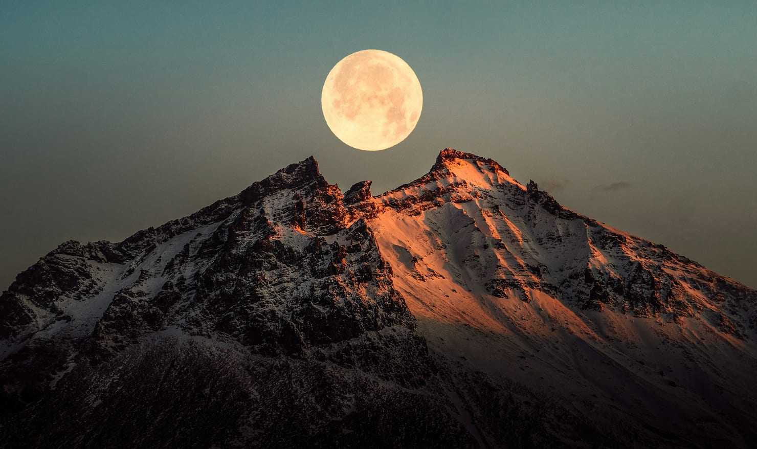 A full moon rising above a sharp, alpenglow-lit snowy peak against a teal dusk sky.