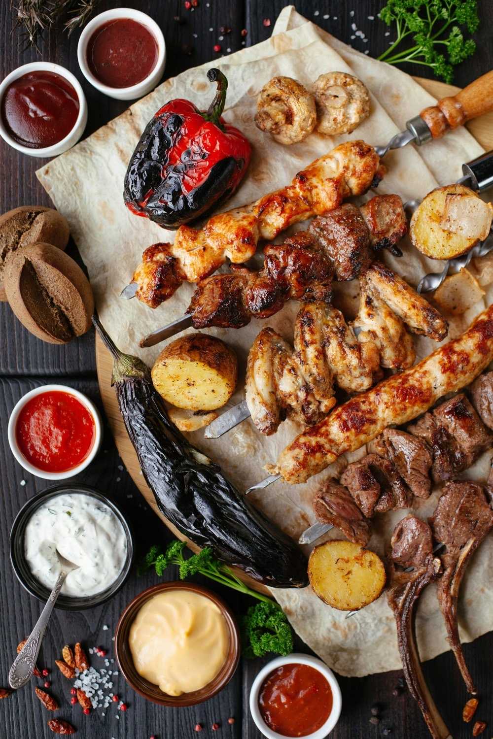 Overhead spread of grilled meat skewers, chicken, roasted pepper, potatoes, and small bowls of dipping sauces on parchment.