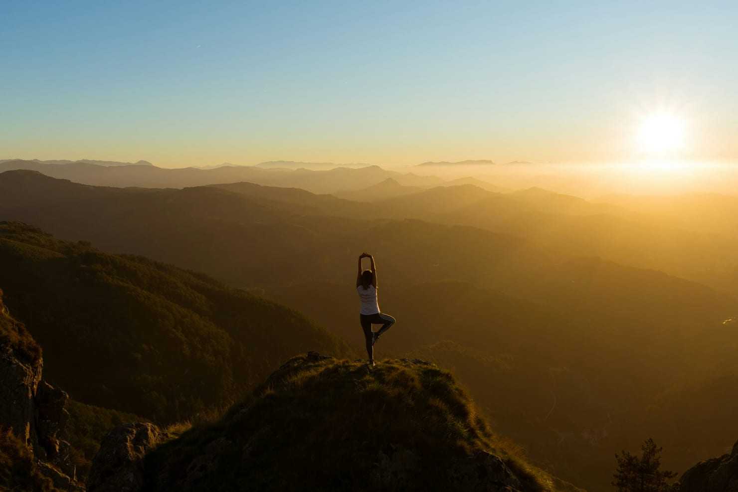 Person in tree pose on a mountain cliff at sunset, golden layered hills behind.