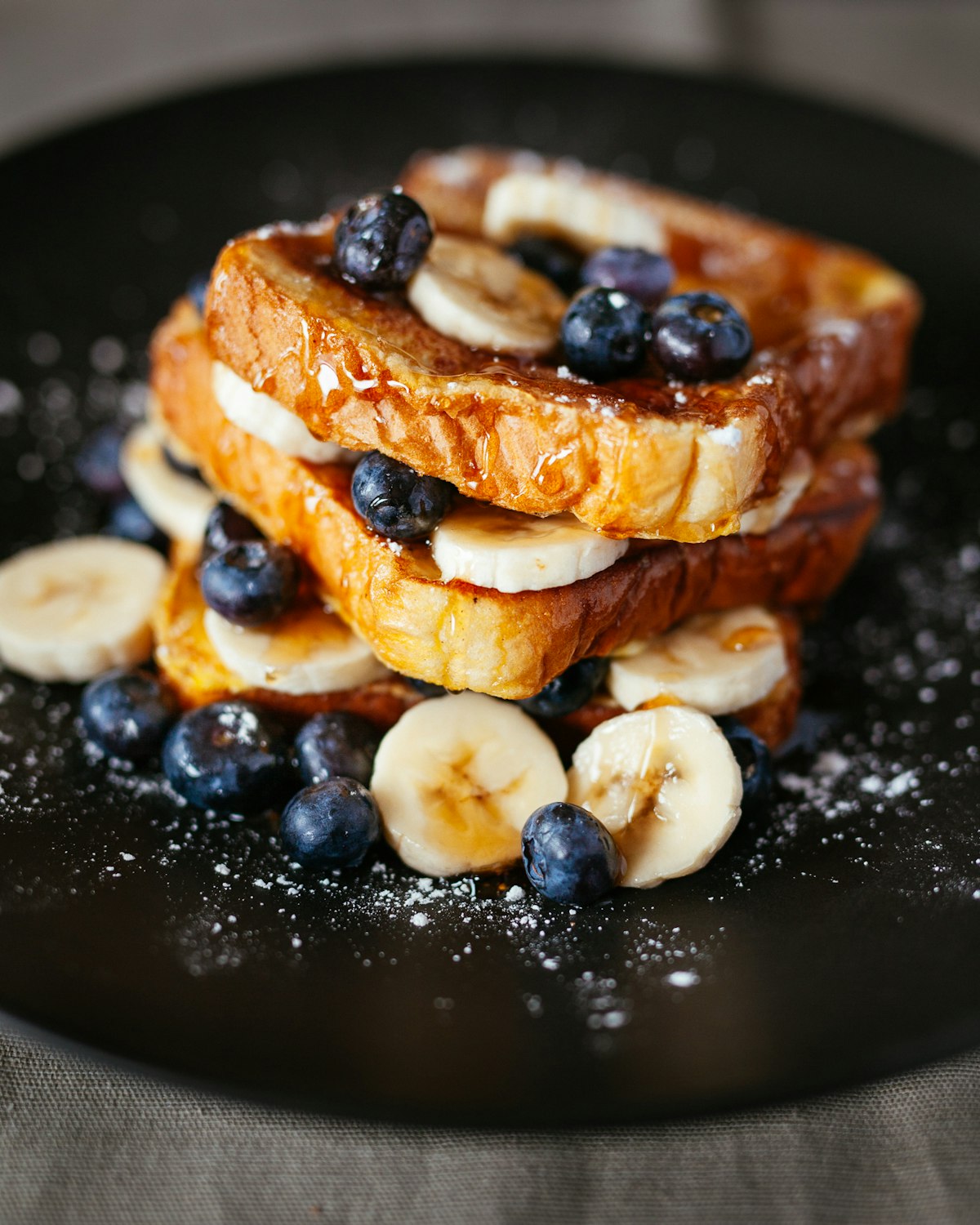A stack of French toast with blueberries, sliced banana, and a dusting of powdered sugar on a black plate.
